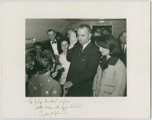 Vice President Lyndon Baines Johnson taking the presidential oath of office surrounded by people in a plane, two hours and eight minutes after the assassination of President John F. Kennedy. Johnson is holding up his right hand, facing U.S. District Judge Sarah T. Hughes, who has her back to the camera.