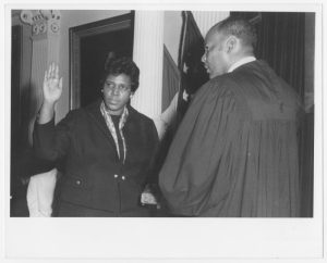 Barbara Jordan being sworn in as Governor for a Day. She is standing and holding her right hand in the air, facing a man with his back to the camera.