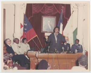 Barbara Jordan standing at a table with microphones, speaking during Governor for a Day. People are sitting watching her. Behind her, there is a portrait and five flags instead of six.