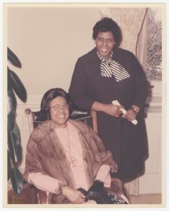 Barbara Jordan standing, leaning on the chair her mother, Arlyne Jordan, is sitting on. They are at the Governor for a Day Ceremony, both smiling.