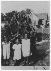 Young Bennie, Barbara, and Rose Mary Jordan standing outside in front of a large bush in light-colored dresses, with a house in the background.