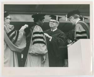 Barbara Jordan wearing a cap and gown with three black stripes on the sleeves, facing a man in a fully black cap and gown. There are two other men on either side of them, wearing the same cap and gown as Barbara Jordan. She is being awarded an honorary Doctor of Law degree at Boston University.