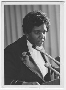 Close-up of Barbara Jordan speaking at a podium in front of microphones. She is wearing a light-colored shirt, a dark jacket, and earrings.