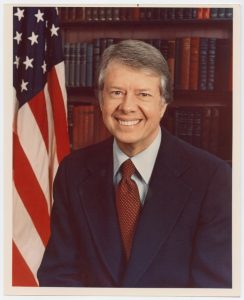 President Jimmy Carter smiling at the camera in a suit, with a bookcase and an American flag in the background.