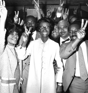 Shirley Chisholm surrounded by members of her campaign team. They are all smiling and giving the V sign with their hands.