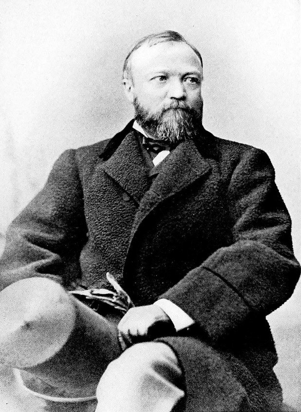 Seated white man with beard wearing large black coat and holding top hat in black and white