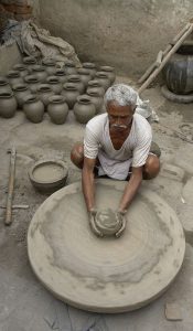 Seated older man in white shirt at wheel on ground, with arrangement of clay pots in background