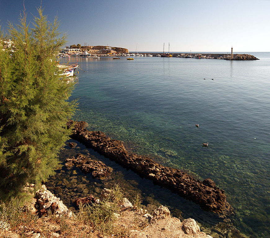 Ruins of Roman port alongside blue green water, with modern buildings and boats in distance