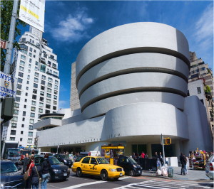 Guggenheim with car-filled street in foreground, including bright yellow taxi. Sunlit with blue sky.