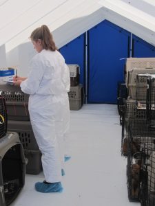 A volunteer wearing a white Tyvek suit and shoe covers writing on a clipboard in an air-conditioned, fully enclosed tent.