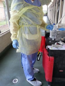 A shelter staff member wearing a loosely-tied yellow gown and white shoe covers. The gown does not cover his leg below the knee, some of the chest and left shoulder are exposed, and the shoe covers do not protect the top of the shoe.