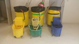 Three large trash cans and three mop buckets. The trash cans are color-coded with yellow, green, or blue tape. The colored mop buckets (yellow, green, blue) are matched to the corresponding trash can color. The color-coded trashcans and mop buckets are assigned to specific housing areas.