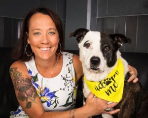 Portrait of Kristen Hassen posing with a black brindle Staffordshire Terrier with a bandana that says “Adopt Me”