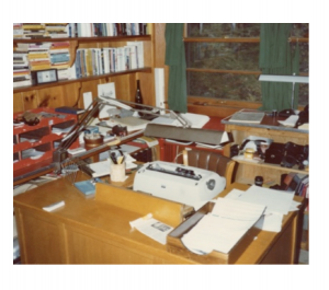 Color photograph of a typewriter and piles of paper on a desk.