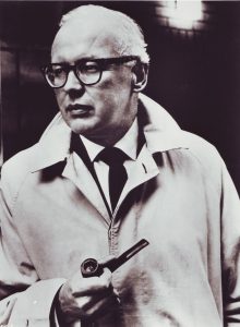 Black and white photograph of MacDonald's "signature look": his pipe, his cigarette lighter, and his trench coat