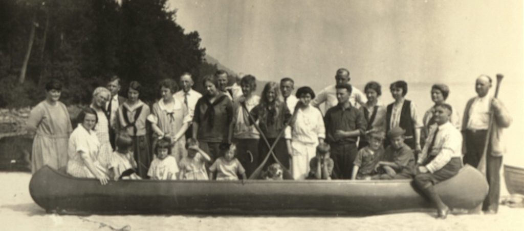Black and white photograph of a group standing behind children in a long canoe. Dorothy stands in the center, smiling.
