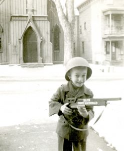 Sepia photograph of a young boy holding a toy gun, dressed in a costume military uniform and hat.